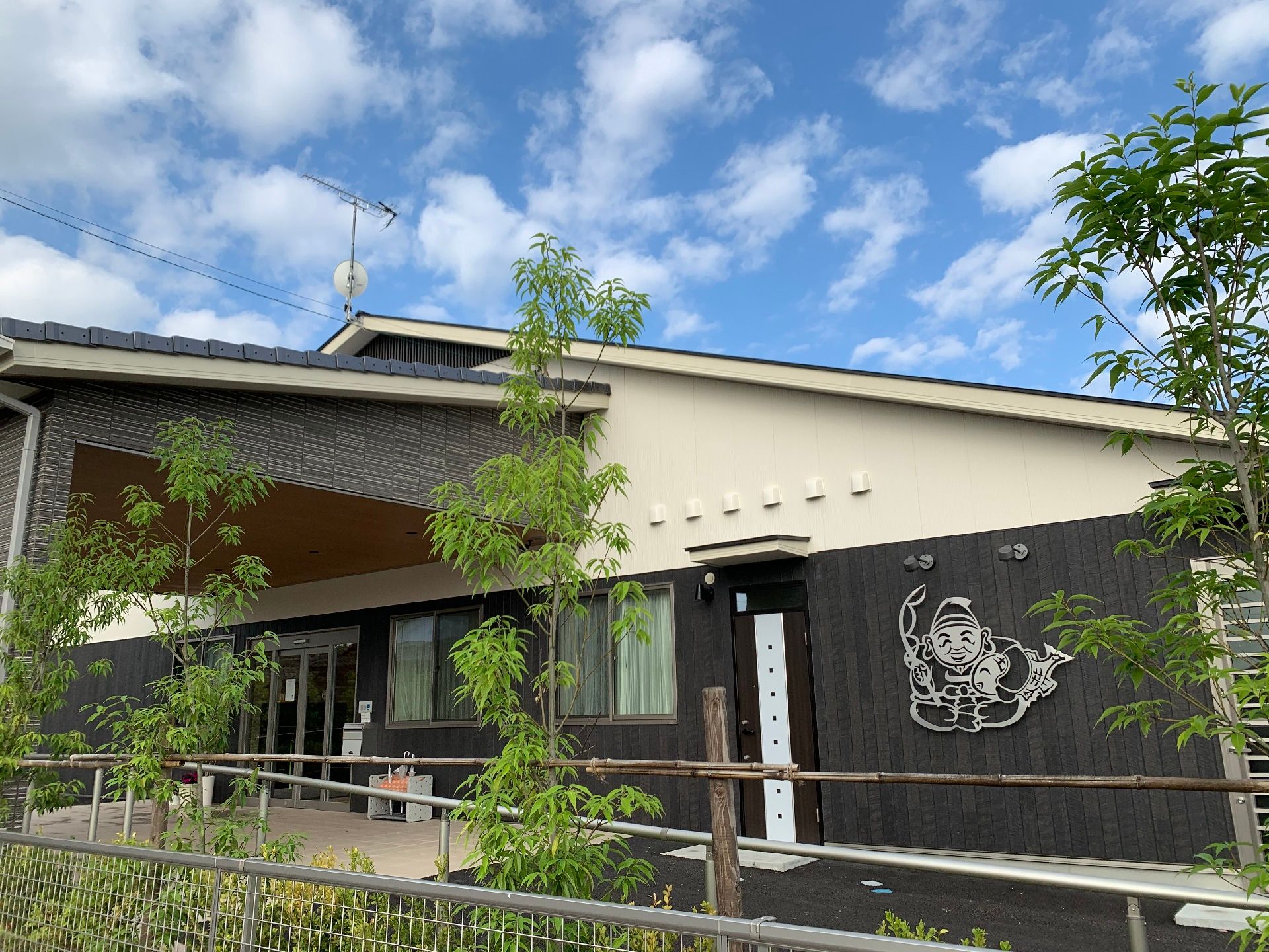 白とグレーの建物は和風の雰囲気があり、グレーの外壁には白いラインで七福神の恵比寿様が描かれている。テラスも付き、周りには緑も植えられている。