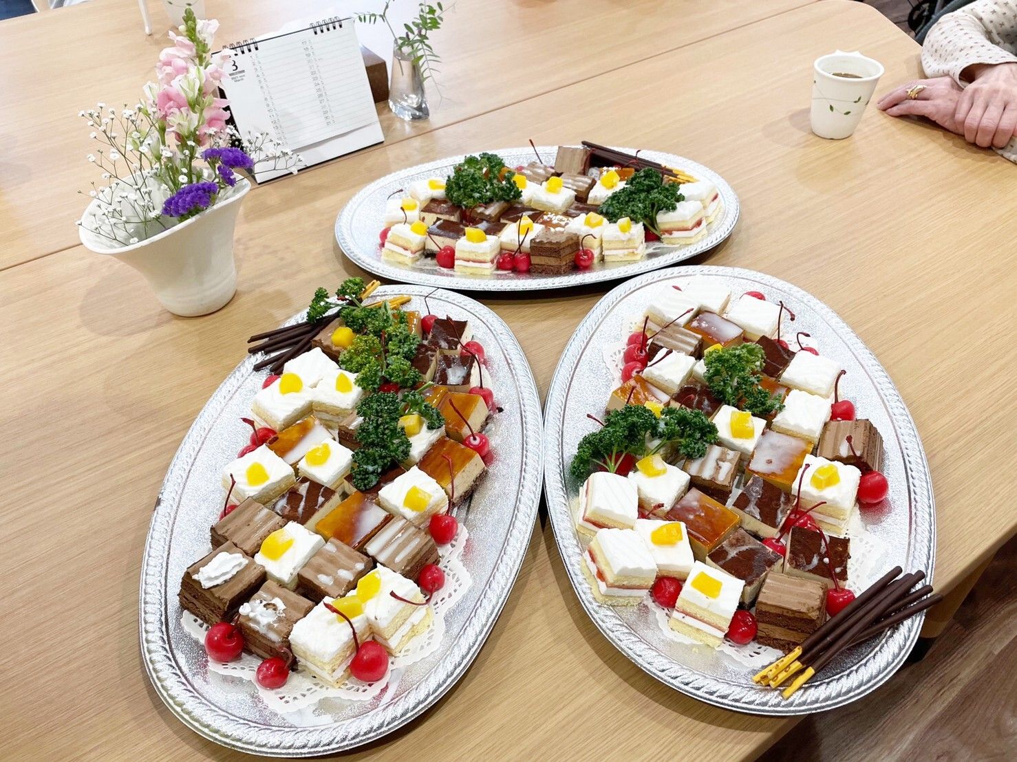 楕円形のシルバーの器の上にはポッキーや一口サイズのケーキが並べられていて、テーブルの上には花が活けられた花瓶も置かれている。