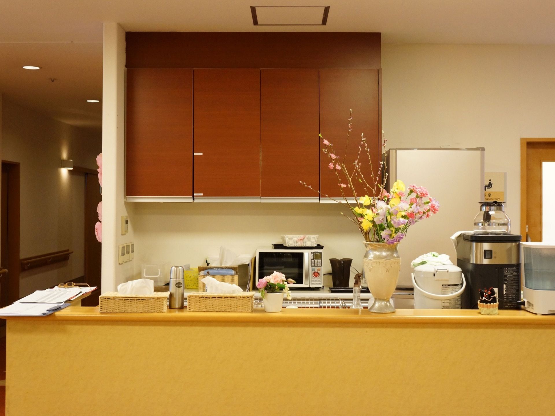 食堂に併設されているキッチンスペース。棚には花が飾られ、冷蔵庫、電子レンジ、コーヒーメーカーやポット等が置かれている。