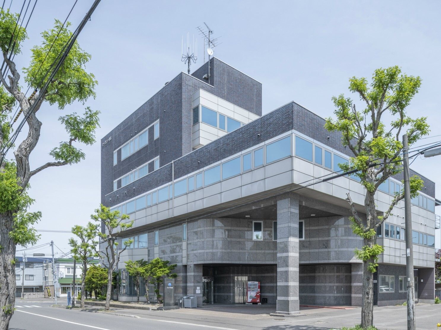 グレー系の建物にはたくさんの窓が付き、周りには木が植えられていて、道路に面している。周りに背の高い建物はない。