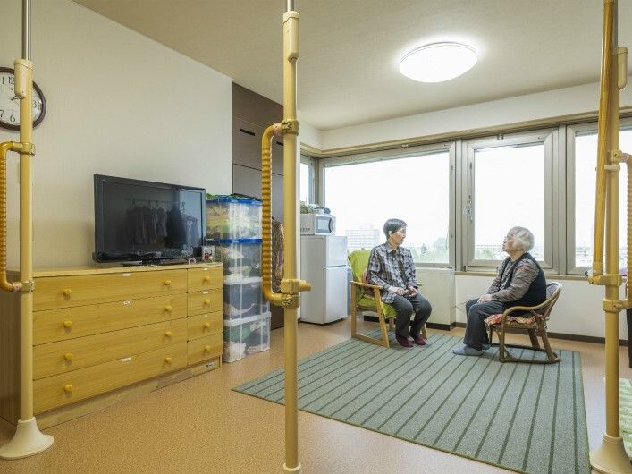 大きな窓が付いた部屋で高齢女性2名がいすに座って話している。部屋の中にはテレビやタンスなども置かれている。