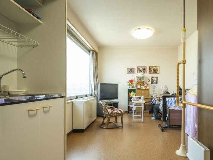 テレビやテーブルが置かれた部屋には絵がたくさん飾られ、ベッドに高齢女性が座っている。手前にはキッチンもある。