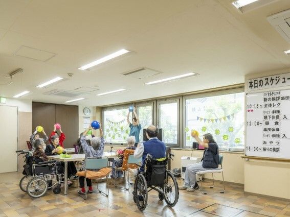明るいホールのような場所で、ボールを使ったエクササイズを行っているスタッフと高齢者たち。車いすの人も一緒にしている。