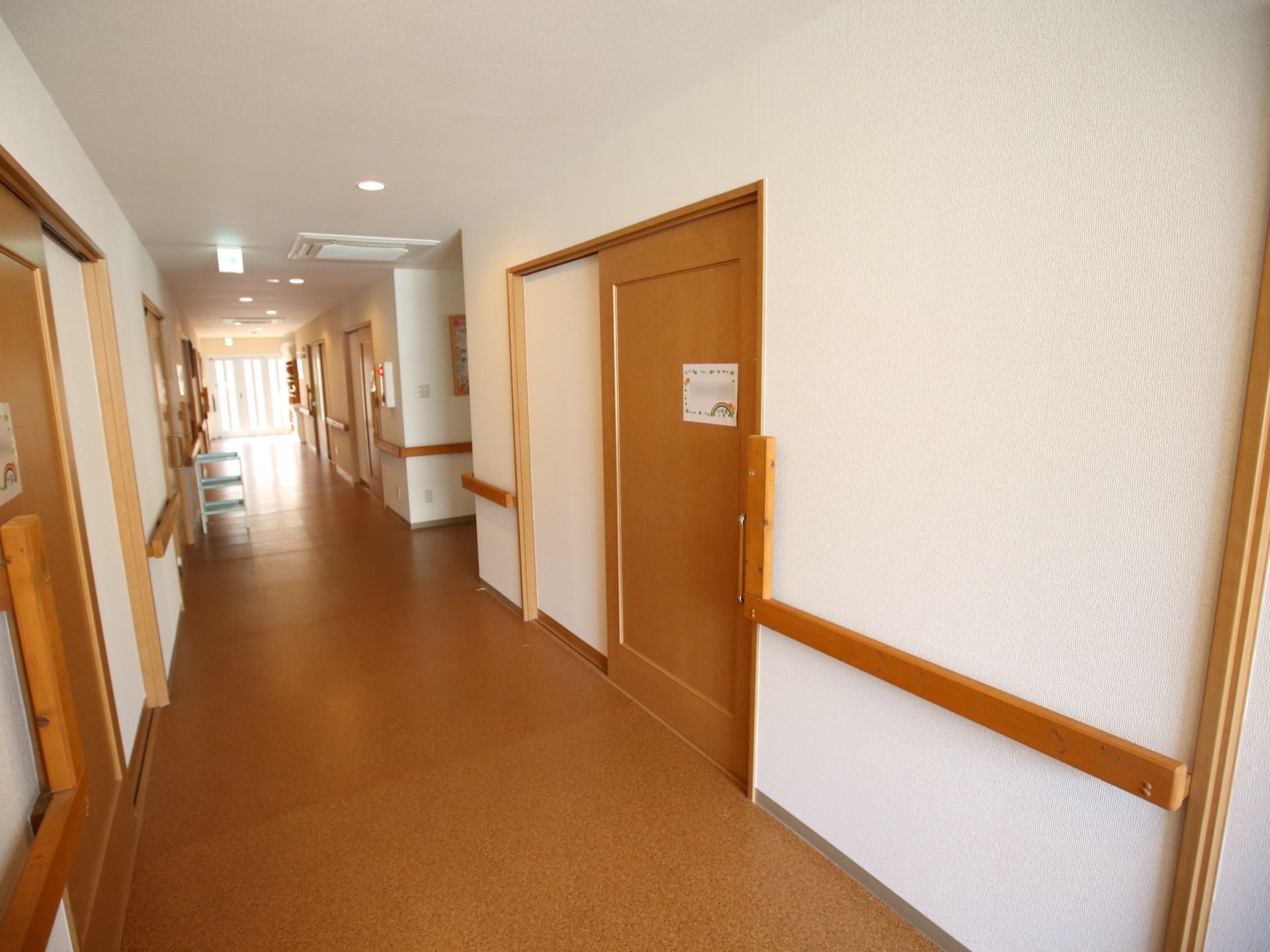 段差がなく、見通しの良い通路である。入居者様の居室が並んでいる。曲がり角でも手すりが途切れず設置され、移動しやすい。