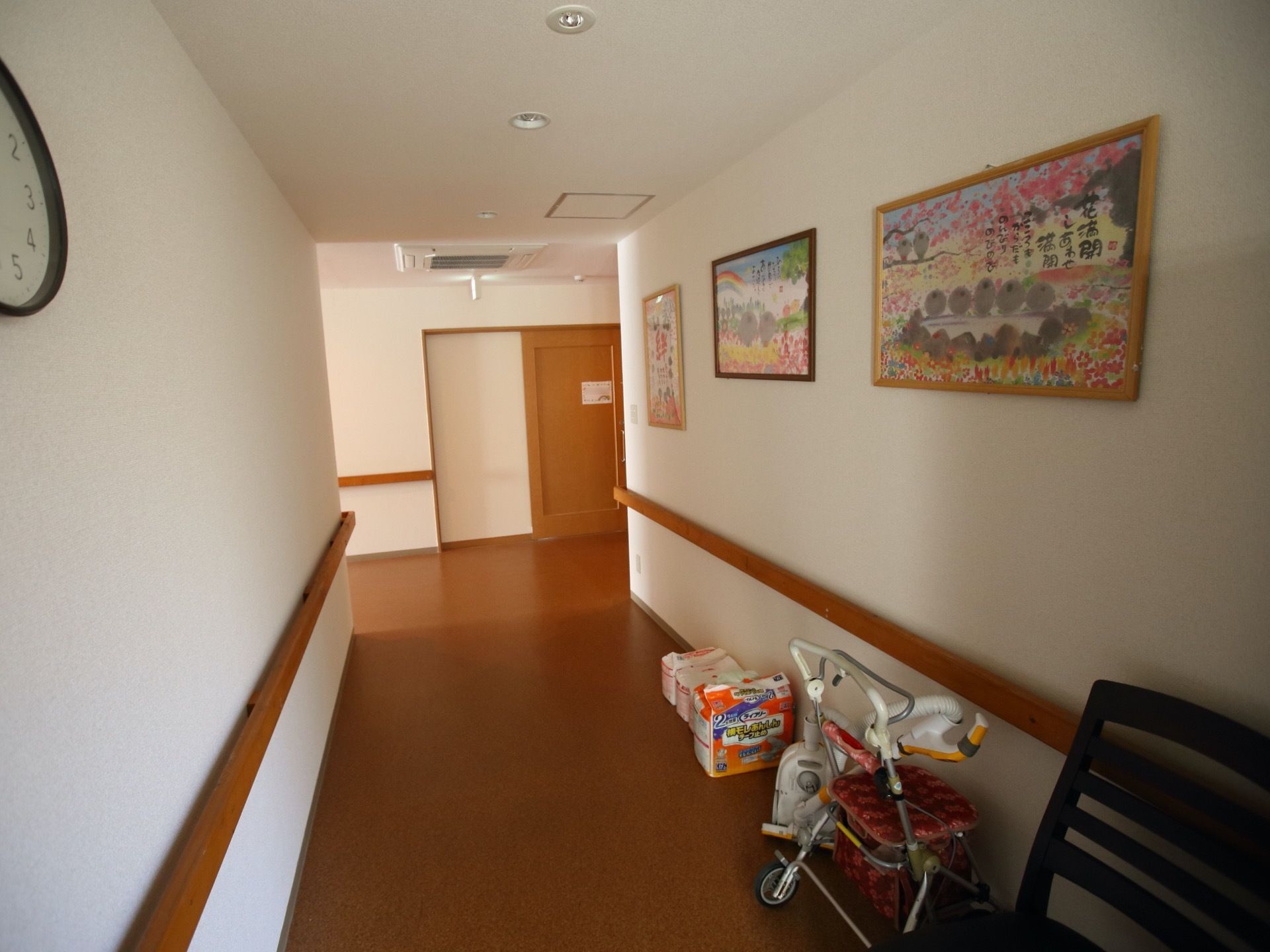段差のないすっきりした通路である。端には、施設の用具が置かれている。白い壁に絵画が飾られ、明るさがある。