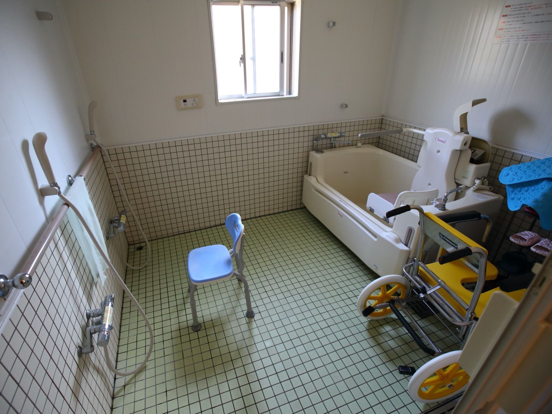 シャワーが２つ並び、シャワーフックの近くに手すりが設置されている。日光が差し込む窓があり、換気がしやすい浴室である。