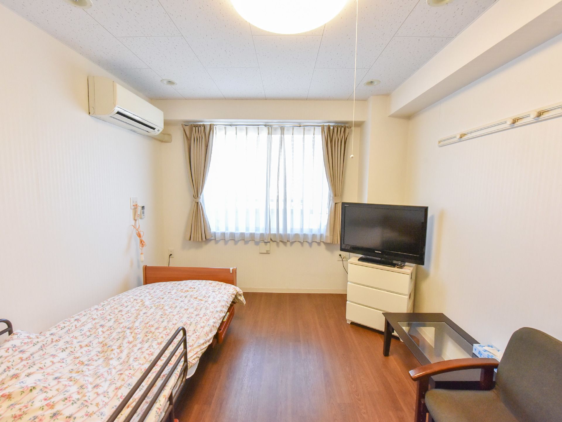 入口側から窓を正面にして居室内を見ると、左手にベッドとエアコン、右手にテレビ、テーブルと椅子がある。右手の壁に壁掛けフックが設置されている。