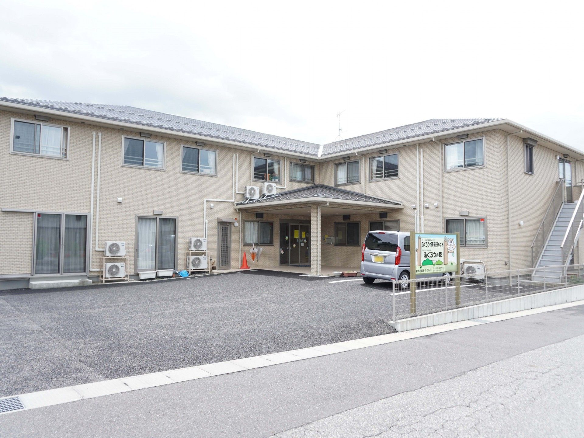 エル字型のブラウンの2階建ての建物には広い駐車場が付き、建物の側面には屋外階段も付いている。