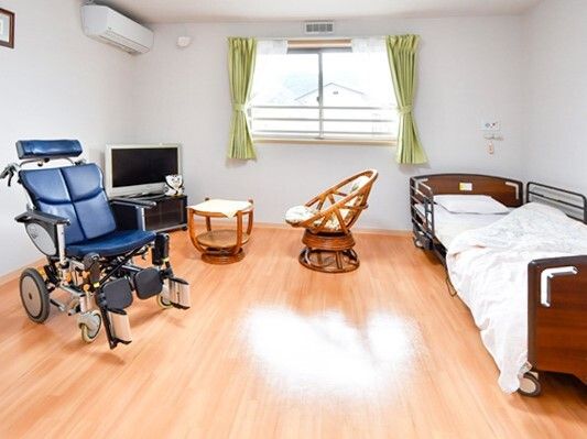 緑のカーテンが付いた大きな窓があるフローリングの部屋にはベッドや車いす、テーブルセットがある。