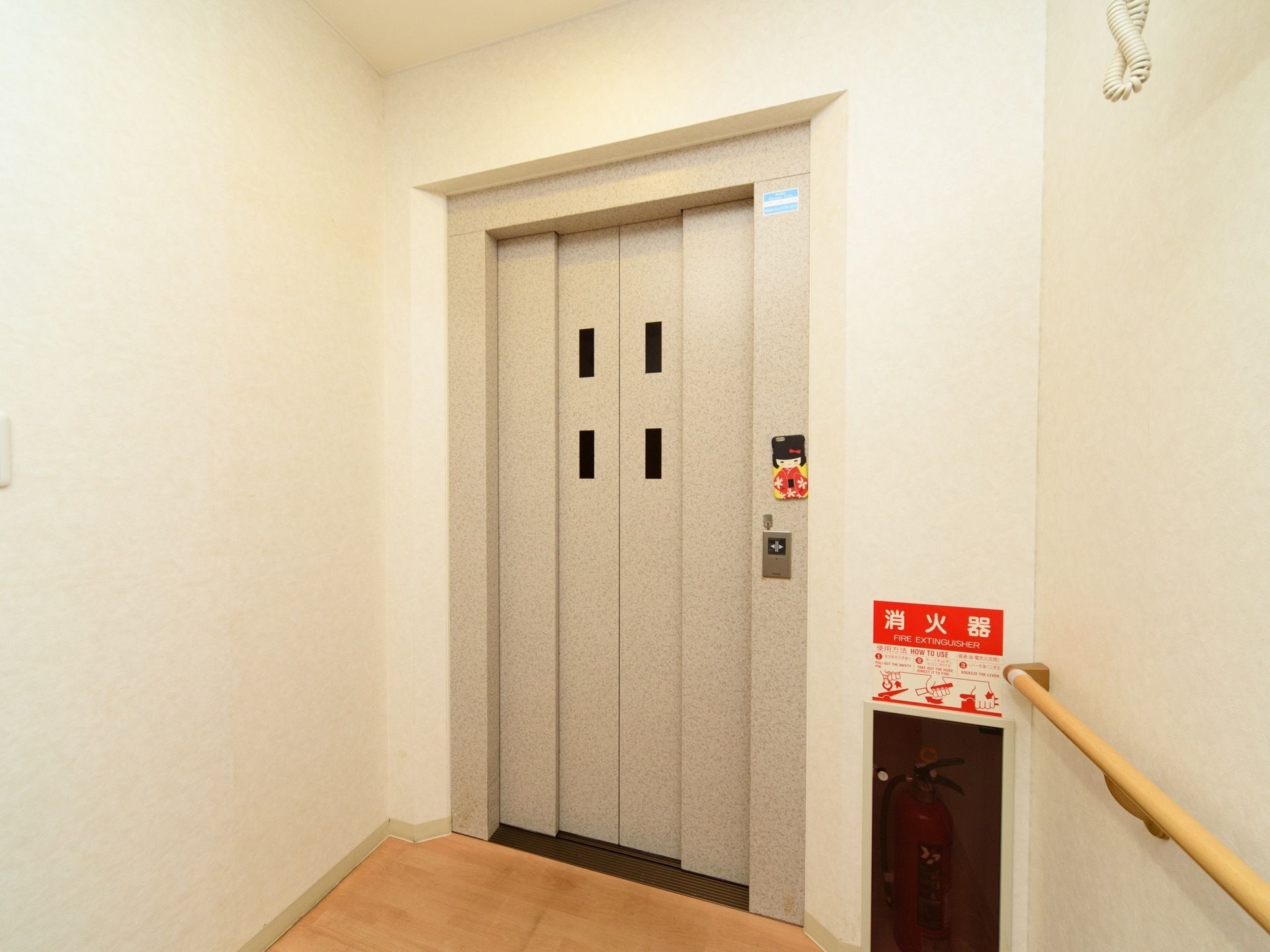 階段の近くにエレベーターが設置されている。壁には手すりが備え付けられており、エレベーターの横には消火器がある。