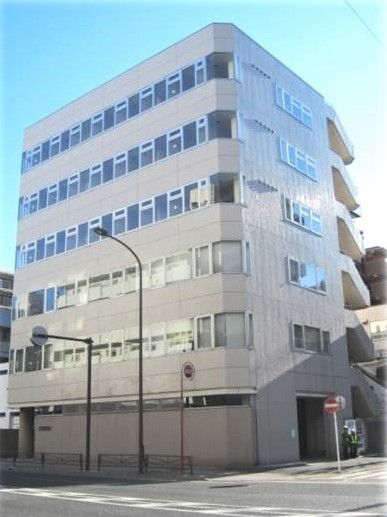 縦に長い６階建ての建物はグレー系で窓が多い。建物の目の前に道路がある。