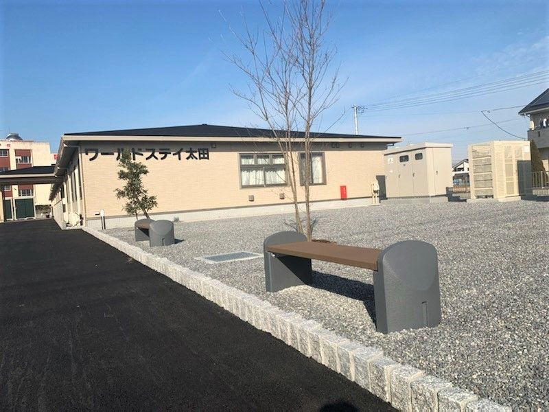 ベージュの平屋建ての建物の外壁にはホーム名が書かれ、ベンチなどが置かれた広いスペースが屋外にある。