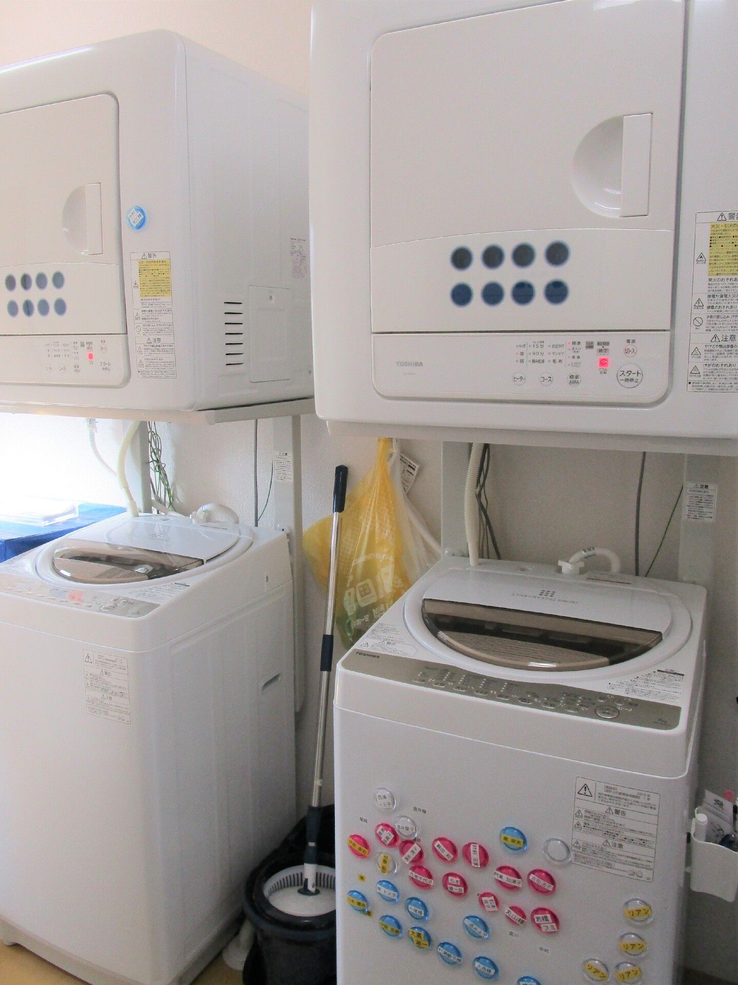 縦型の洗濯機が2台あり、その上にドラム型の乾燥機が2台設置され、洗濯機の一つには赤や青の丸いシールのようなものが貼られている。