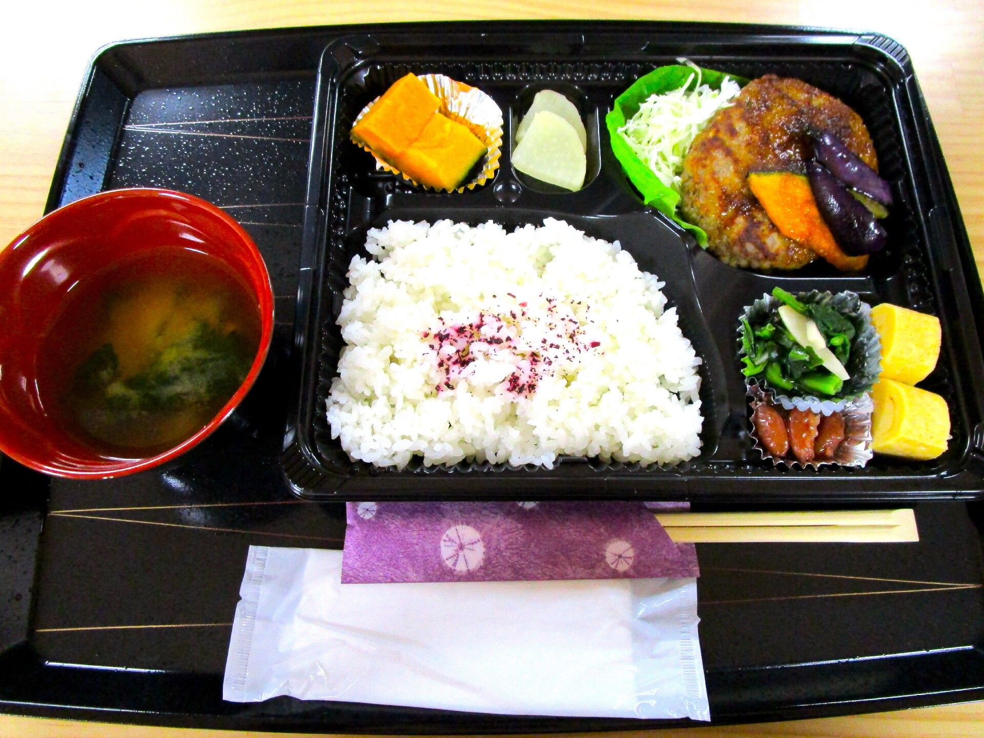 ゆかりご飯に味噌汁、ハンバーグやカボチャの煮物、出汁巻き卵、漬物など、おかずがちょっとずつ盛られている弁当風の食器。