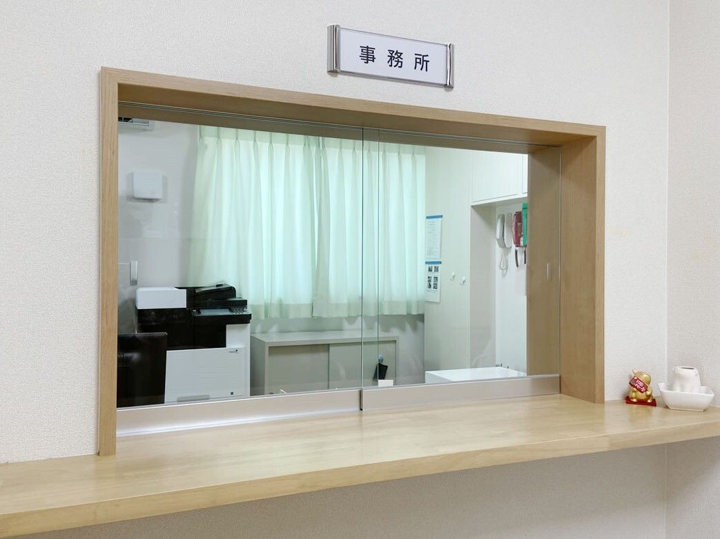 「事務所」と書かれたプレートが貼られた窓の内側には複合機などが置かれている。