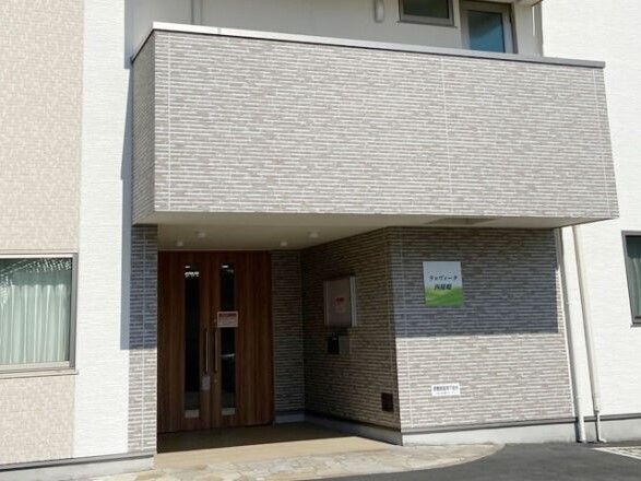 グレーの建物の玄関には住宅名が書かれたプレートが貼られ、2階部分のベランダが玄関の屋根となっている。