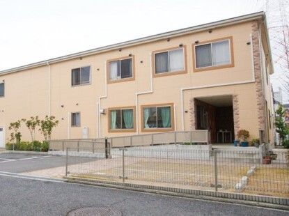 駐車場付きのベージュの2階建ての建物。住宅地の中にあり、圧迫感がなく親しみやすく見える外観。