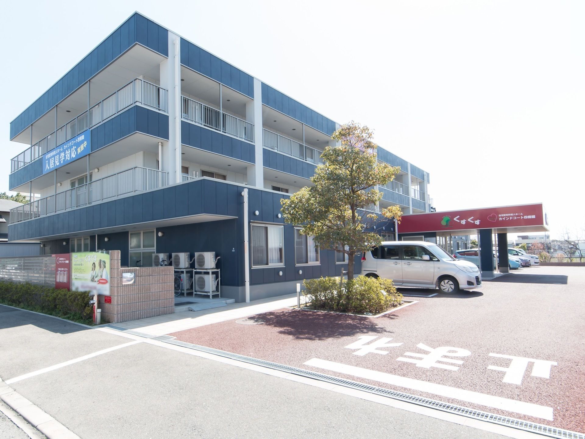 敷地内に広い駐車場が完備されている住まいで、複数の車が停められている。玄関の屋根部分には、施設名が書かれている。