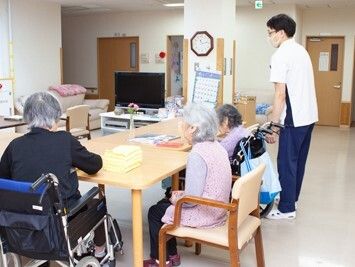 ３名の高齢者がテーブルを囲んでいる。2名は車いすの利用者で、一人には男性スタッフが付き介助をしている。