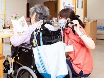 本を読んでいる車いすの高齢女性のそばにはオレンジのポロシャツを着た女性スタッフがいて見守っている。