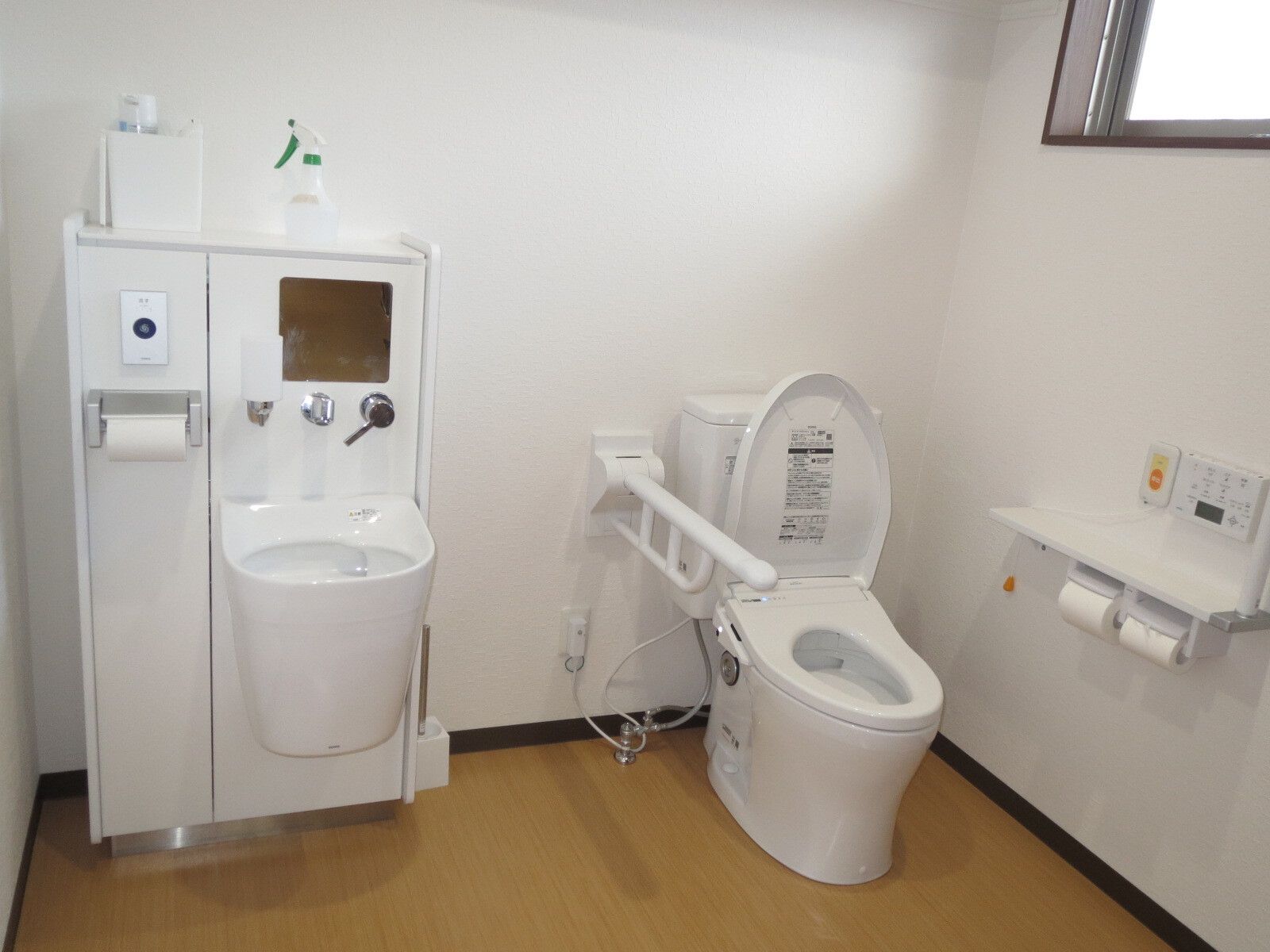 オレンジの呼び出しボタンやトイレットペーパーホルダーが設置された壁は白く、トイレは広い。手洗い場もあり、手すりも付いている。