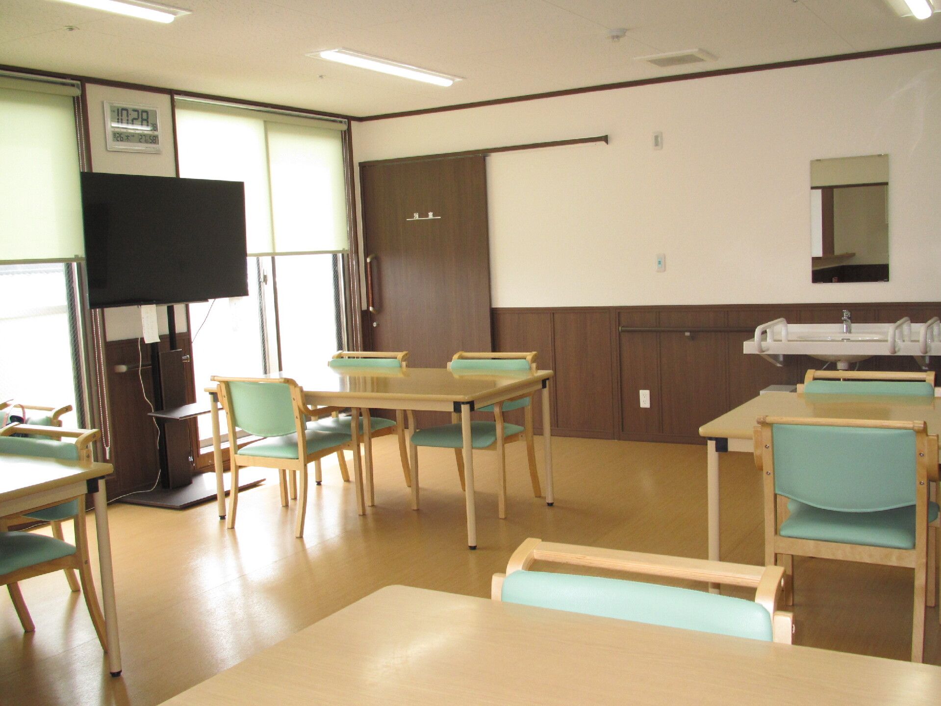 緑の椅子がセットされたダイニングテーブルが複数置かれた食堂には大きなテレビもある。窓にはスクリーンカーテンが付いている。