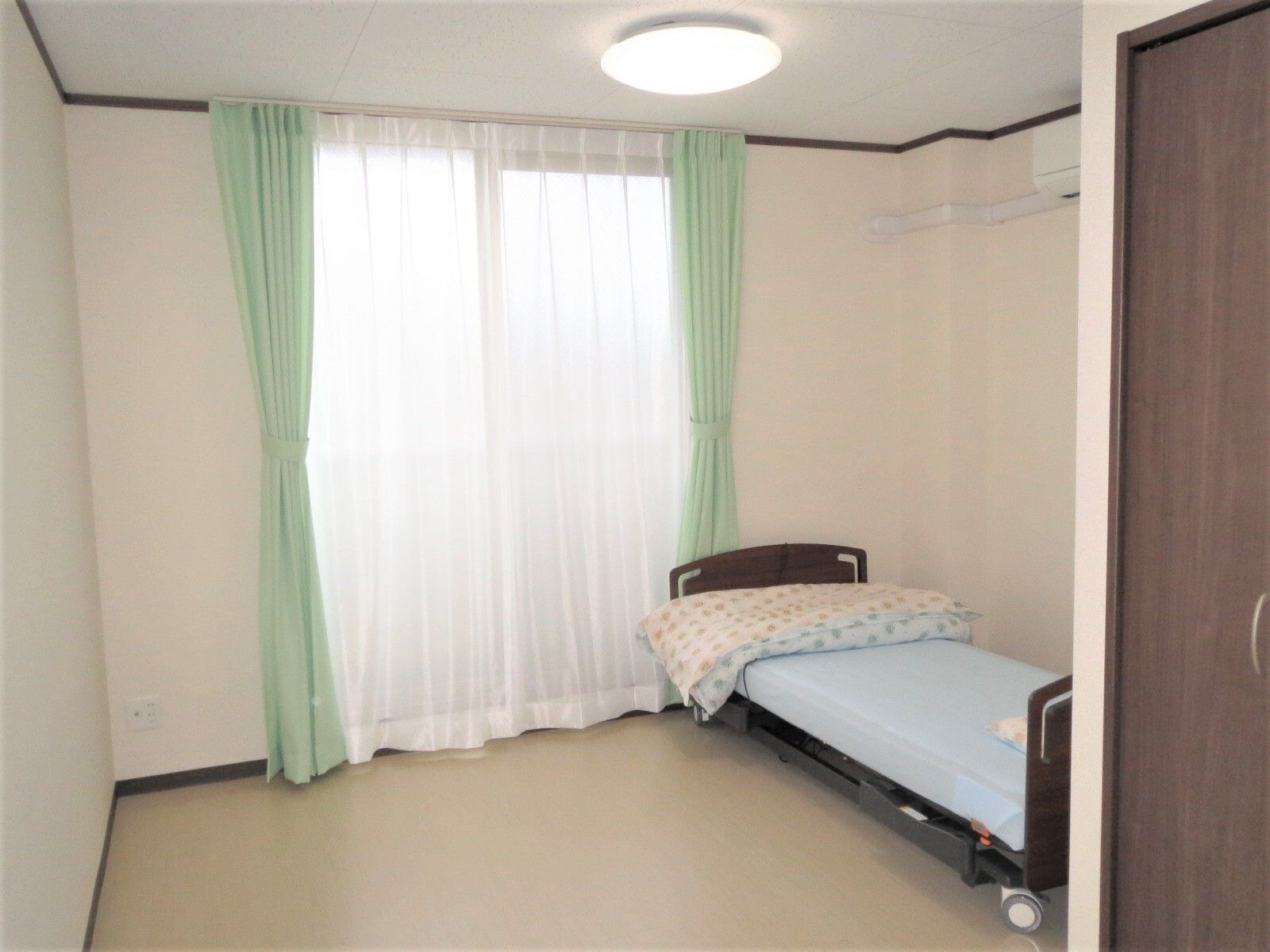 寝具付きのベッドやカーテン付きの窓がある一人用の部屋。壁にはエアコンもついている。手前にはブラウンのドアもある。