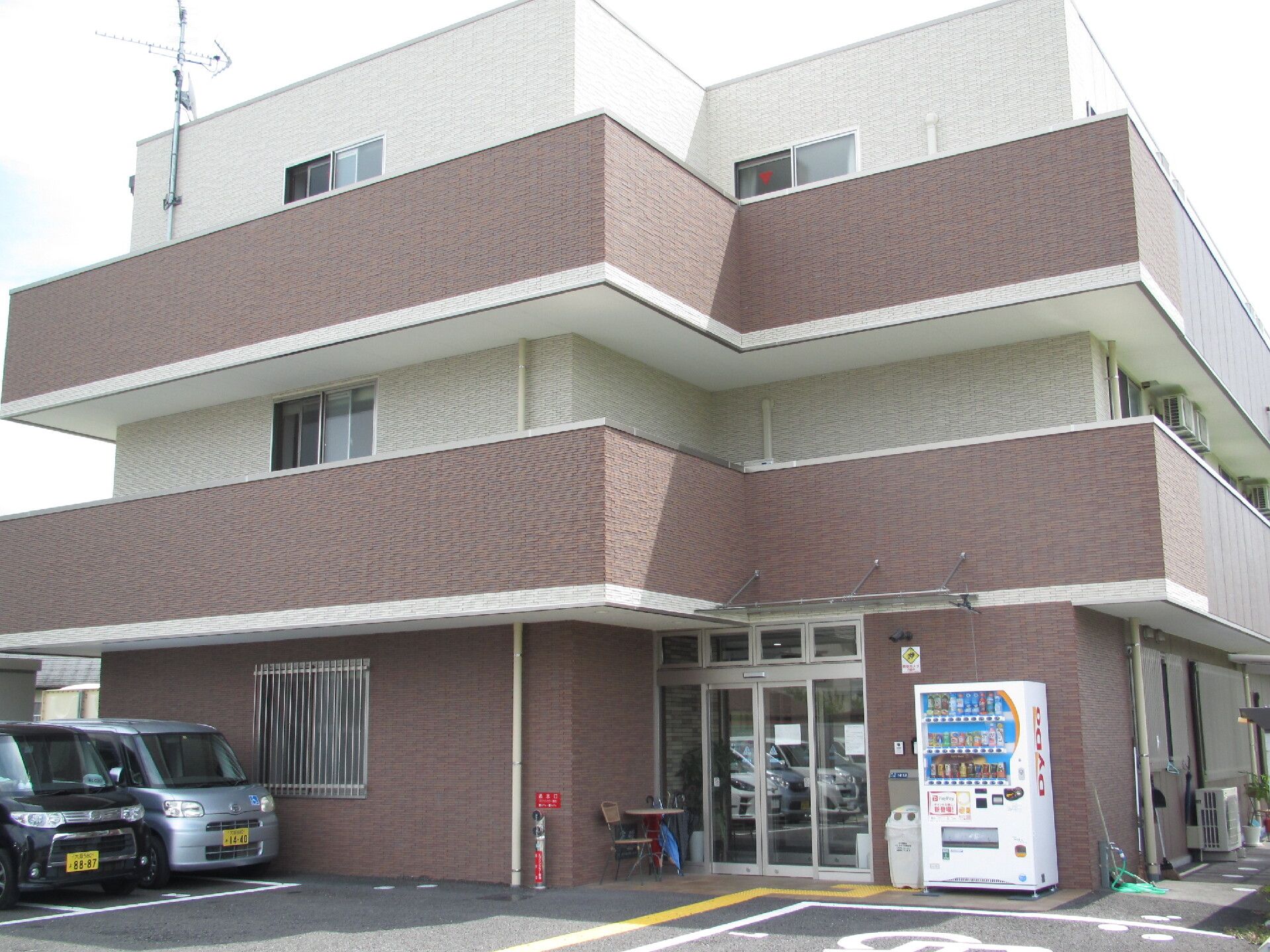 ブラウンと白の3階建ての建物は各フロアにベランダがあり、敷地内には駐車場も完備され、自動販売機も置かれている。