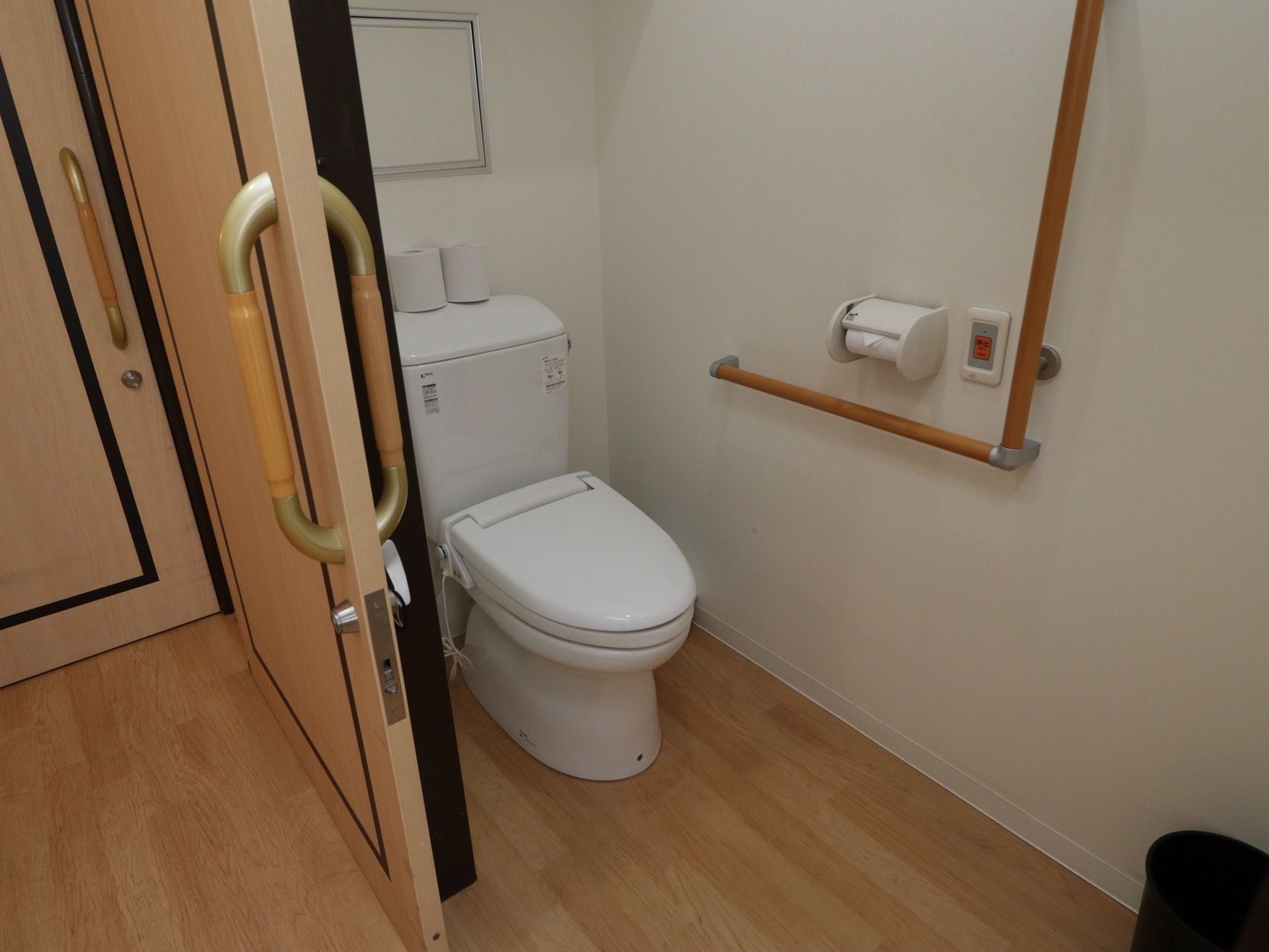 居室のトイレである。Ｌ型手すりや呼出ボタンが完備されており、ゴミ箱が置かれている。扉には、スライドドアが採用されている。