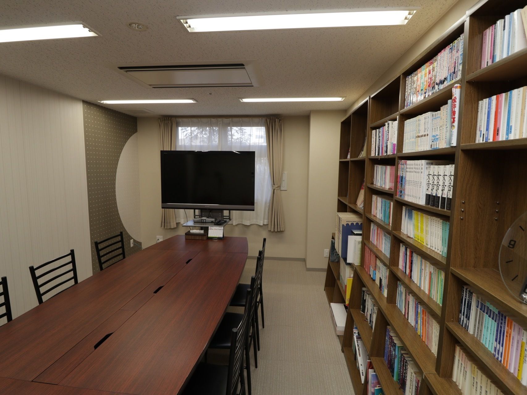 落ち着いた雰囲気の図書室である。中央に大きなテーブルが置かれている。壁際に本棚が設置されており、大型テレビが窓際に置かれている。