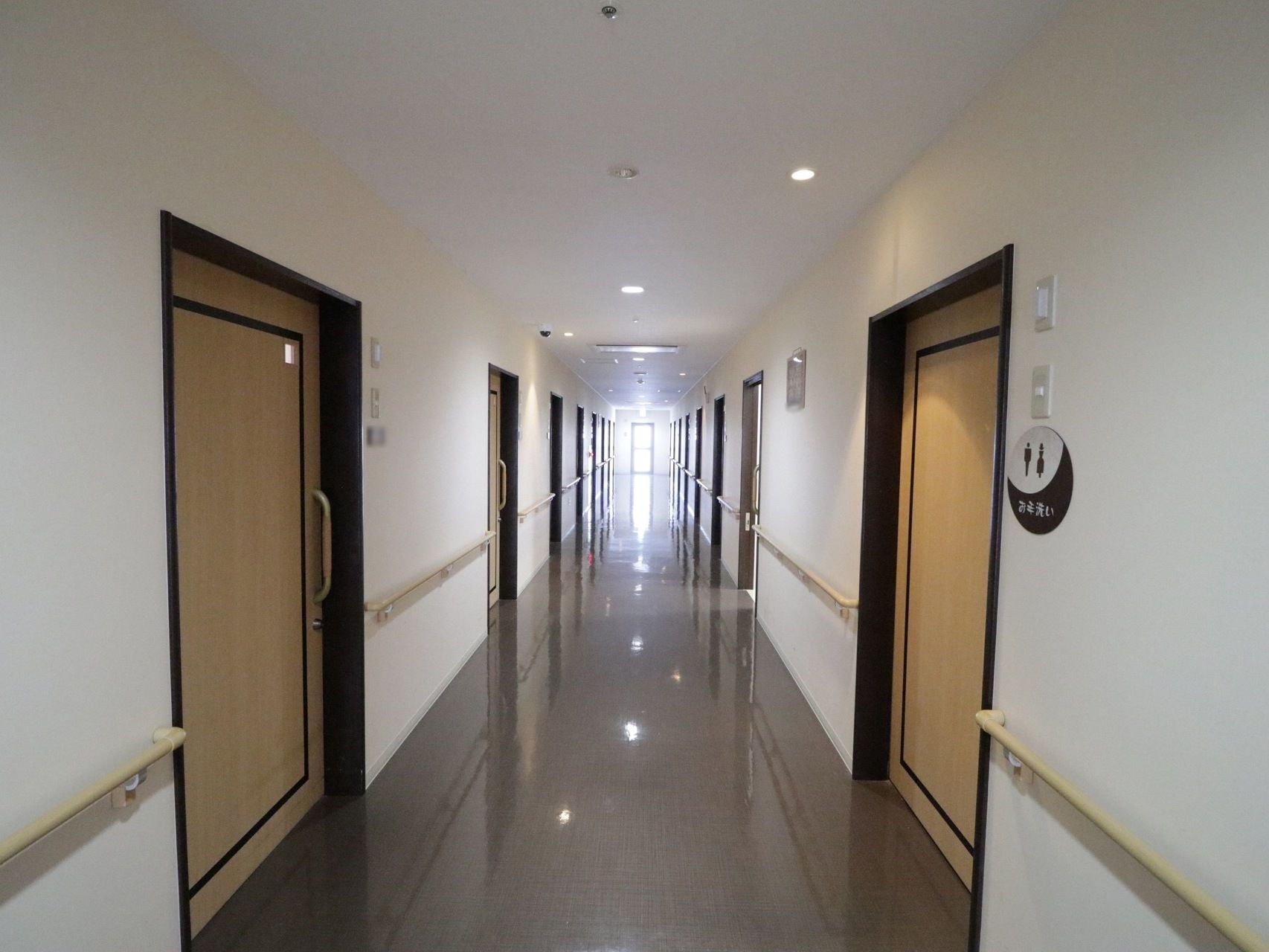 広々とした見通しの良い廊下である。両サイドには、居室やトイレの扉が並んでいる。手すりが壁に沿って取りつけられている。