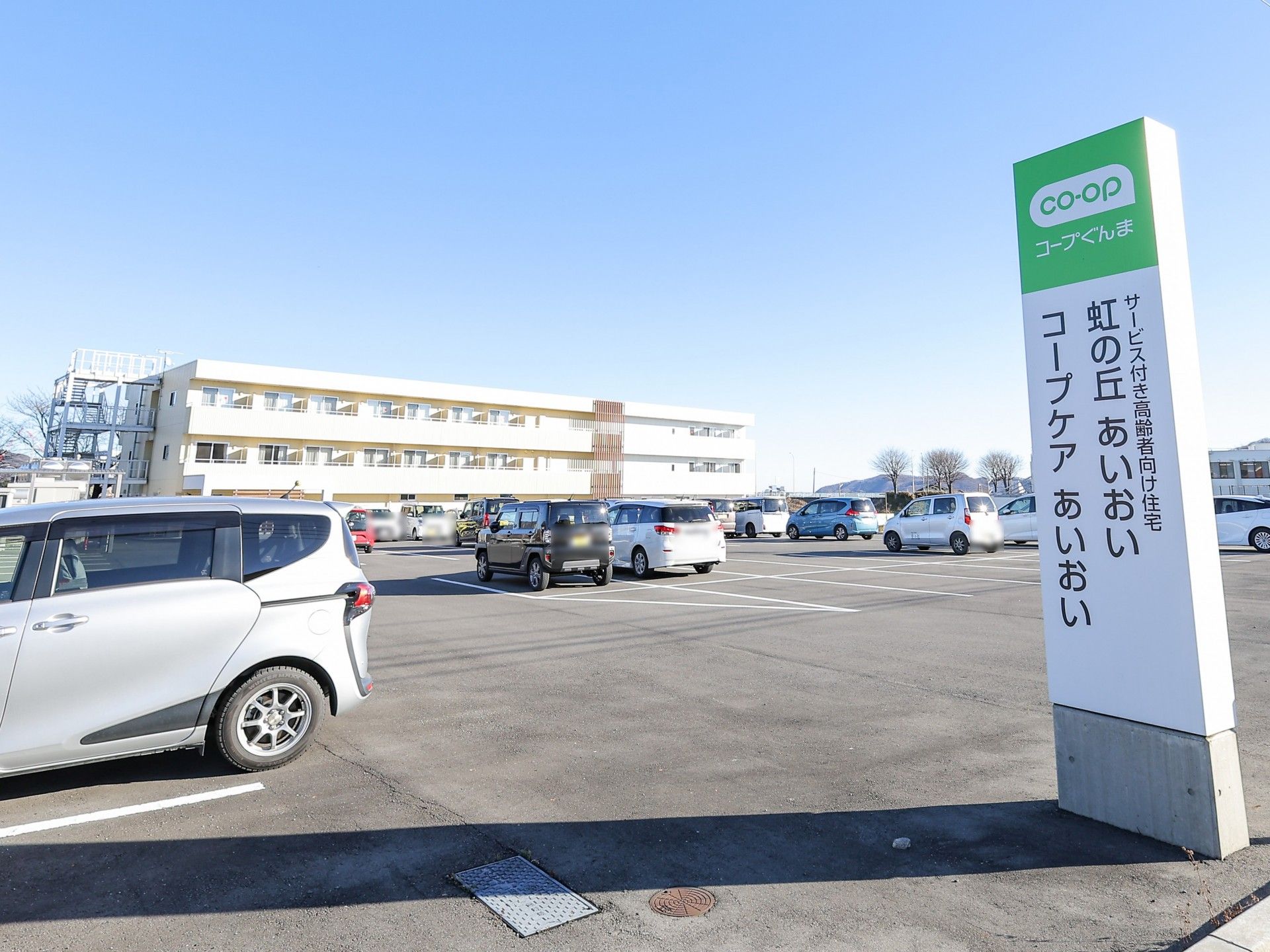 ３階建ての建物は白とベージュで周辺には緑が多くあり、外壁には住宅名が書かれている。