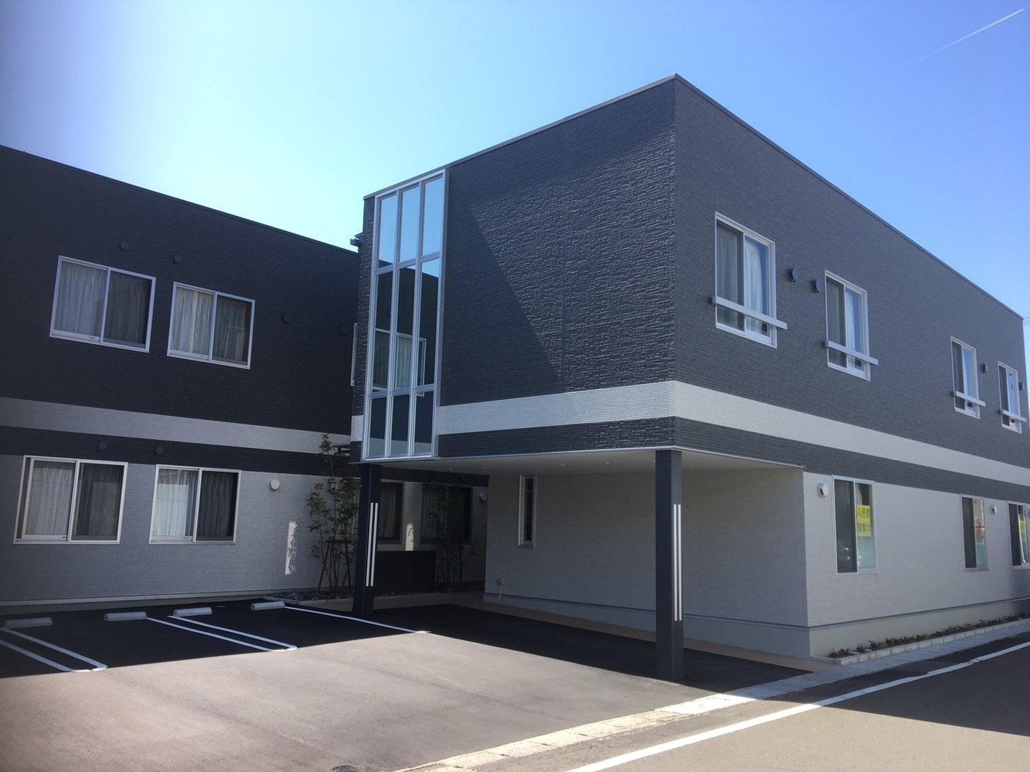 2階建ての建物は横に長く駐車場もある。敷地内には車が止まり、木も植えれられている。
