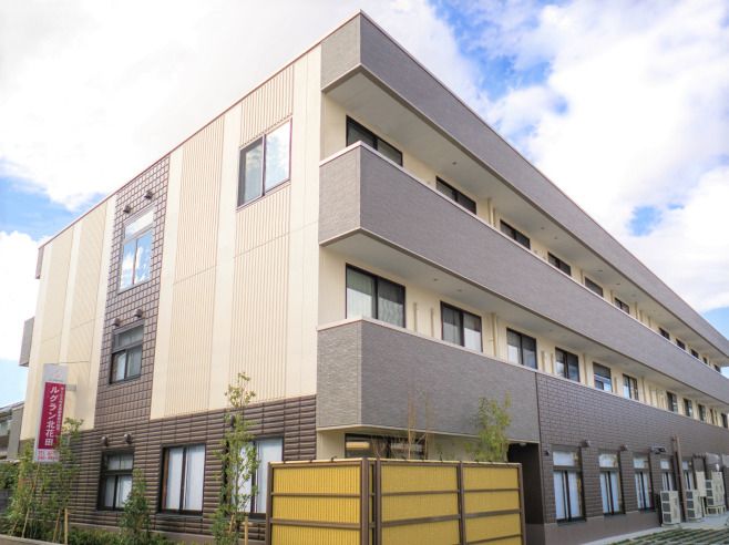 ベランダが各フロアについた3階建ての建物はブラウン系。建物の周辺には木が植えられ、日当たり良好。