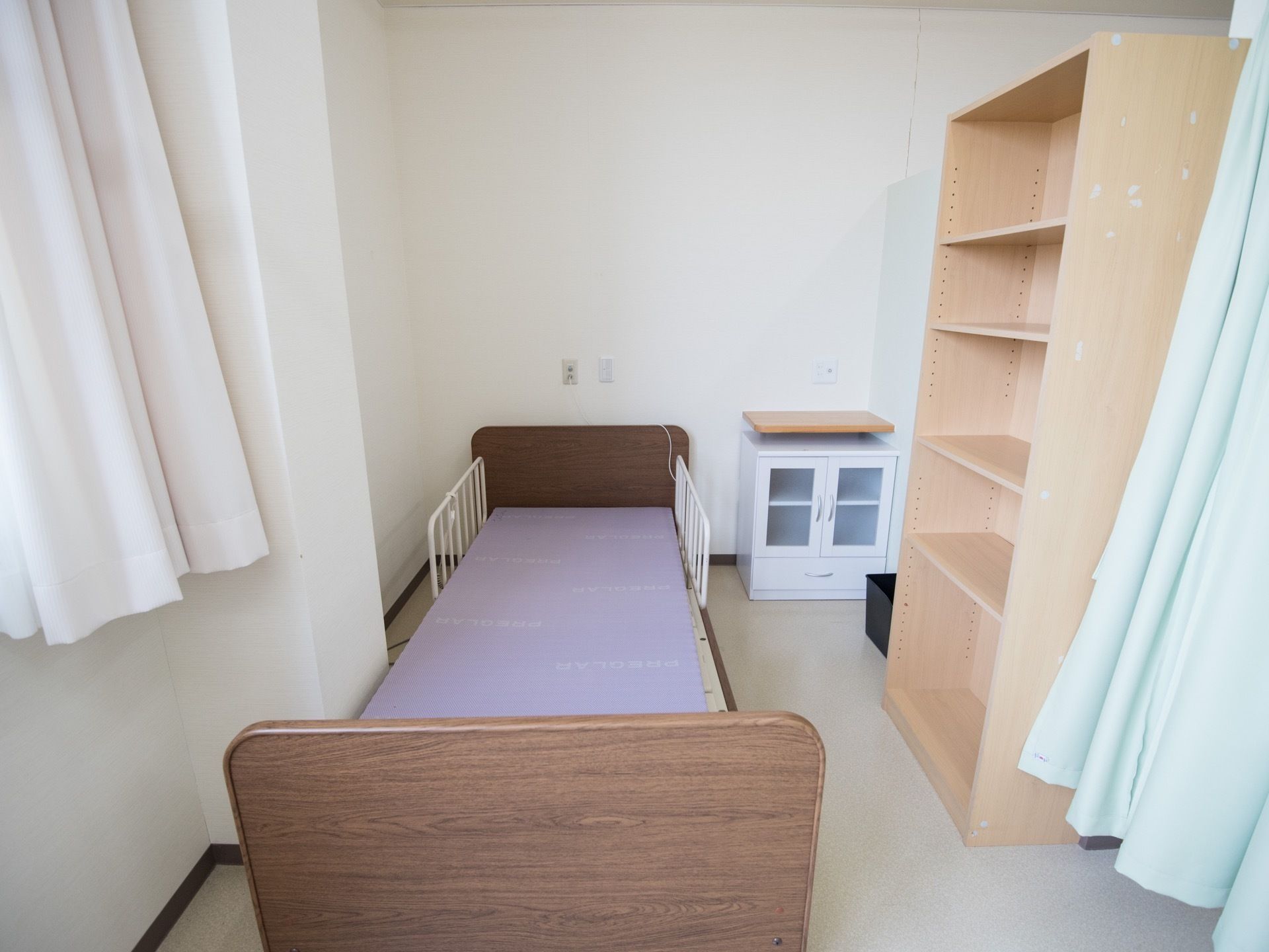 ご入居前の居室の様子である。介護用ベッドが設置され、明るい木目調の仕切り板付き収納棚や白い整理棚が置かれている。