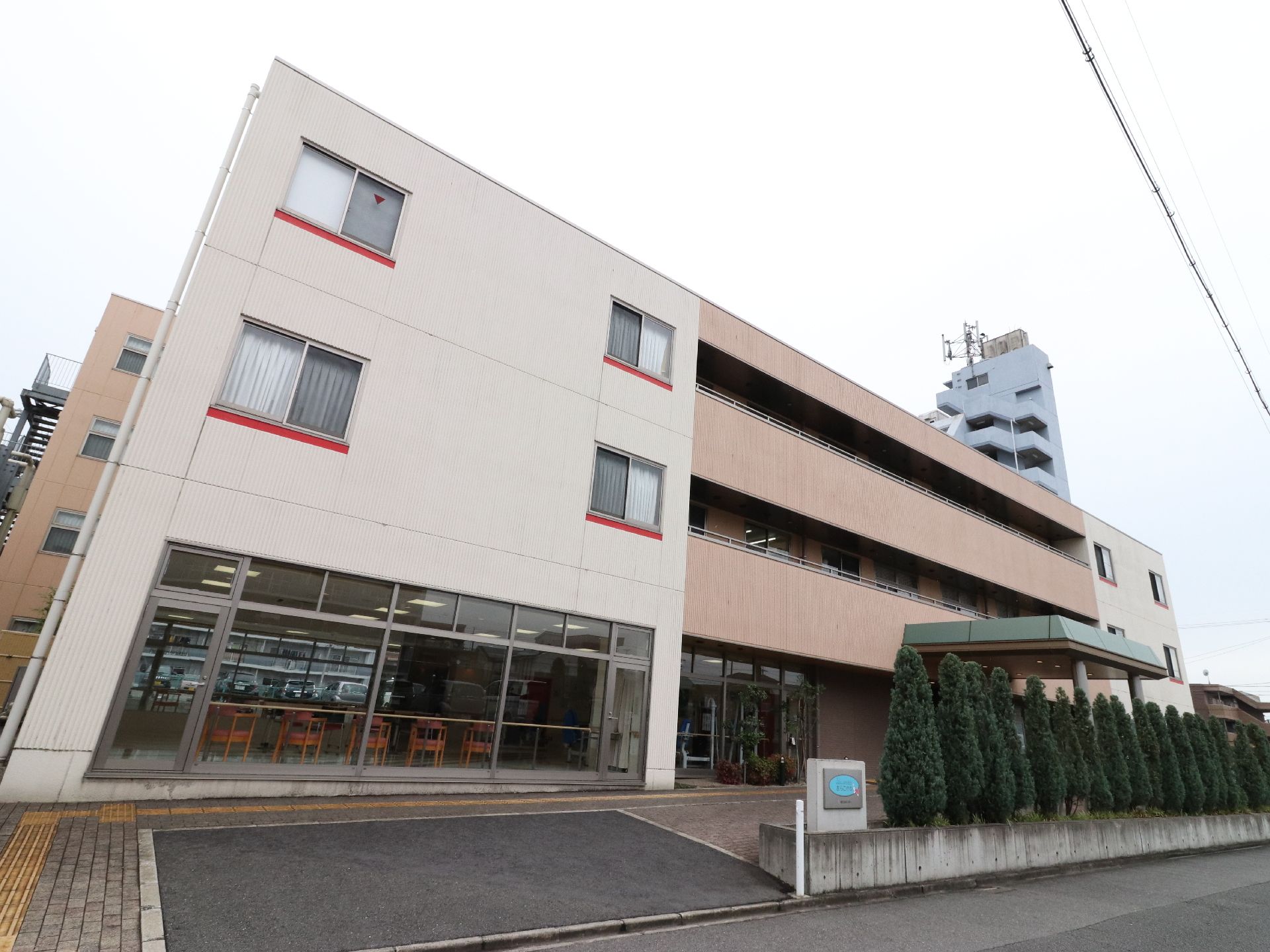 施設外観の様子。白と茶色で長方形の建物。1階の左側は前面がガラス張りとなっていて、中の様子がうかがえる。
