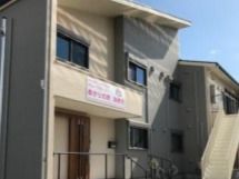 ベージュとグレー系の外観の２階建ての建物は日当たりよく、玄関には屋根があり、ホーム名が書かれた垂れ幕もある。