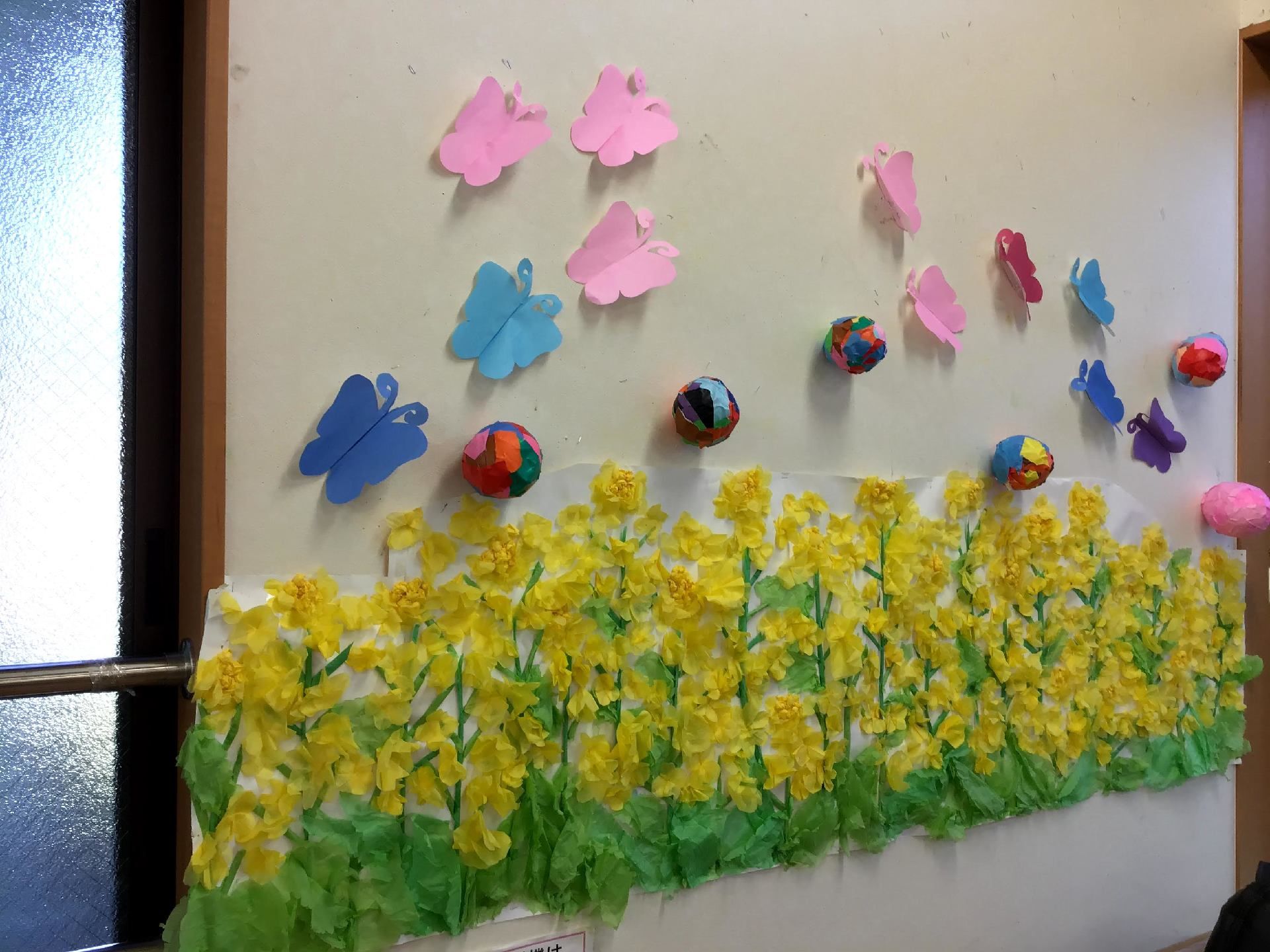 色画用紙で作られた菜の花と蝶々が飾られた白い壁。