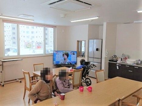 テレビを見ている車いすの入居者やテーブルについている女性入居者がいて、キッチンや冷蔵庫もある。