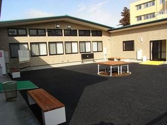 ベンチやテーブル、ゴミ箱などが置かれた中庭が中央にあり、周りを囲むようにブラウン系の外壁の平屋建ての建物がある。