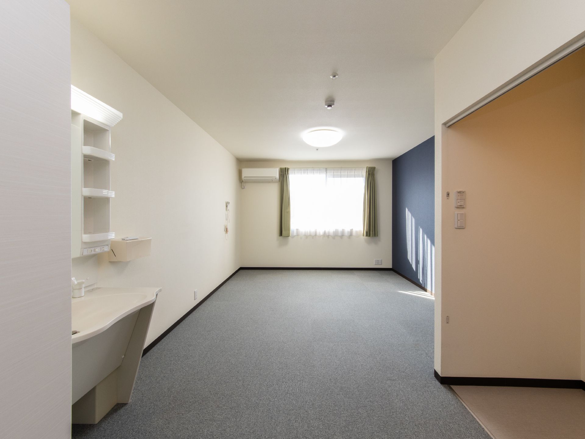 奥行きのある、洗練された内装の居室。奧の壁には窓やエアコンがあり、左側の手前には洗面台が取りつけられている。