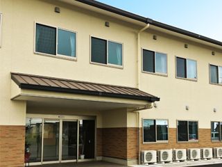 2階建ての建物はベージュとブラウンのレンガ調の外壁でまとめられ、落ち着いた雰囲気。玄関には屋根がある。