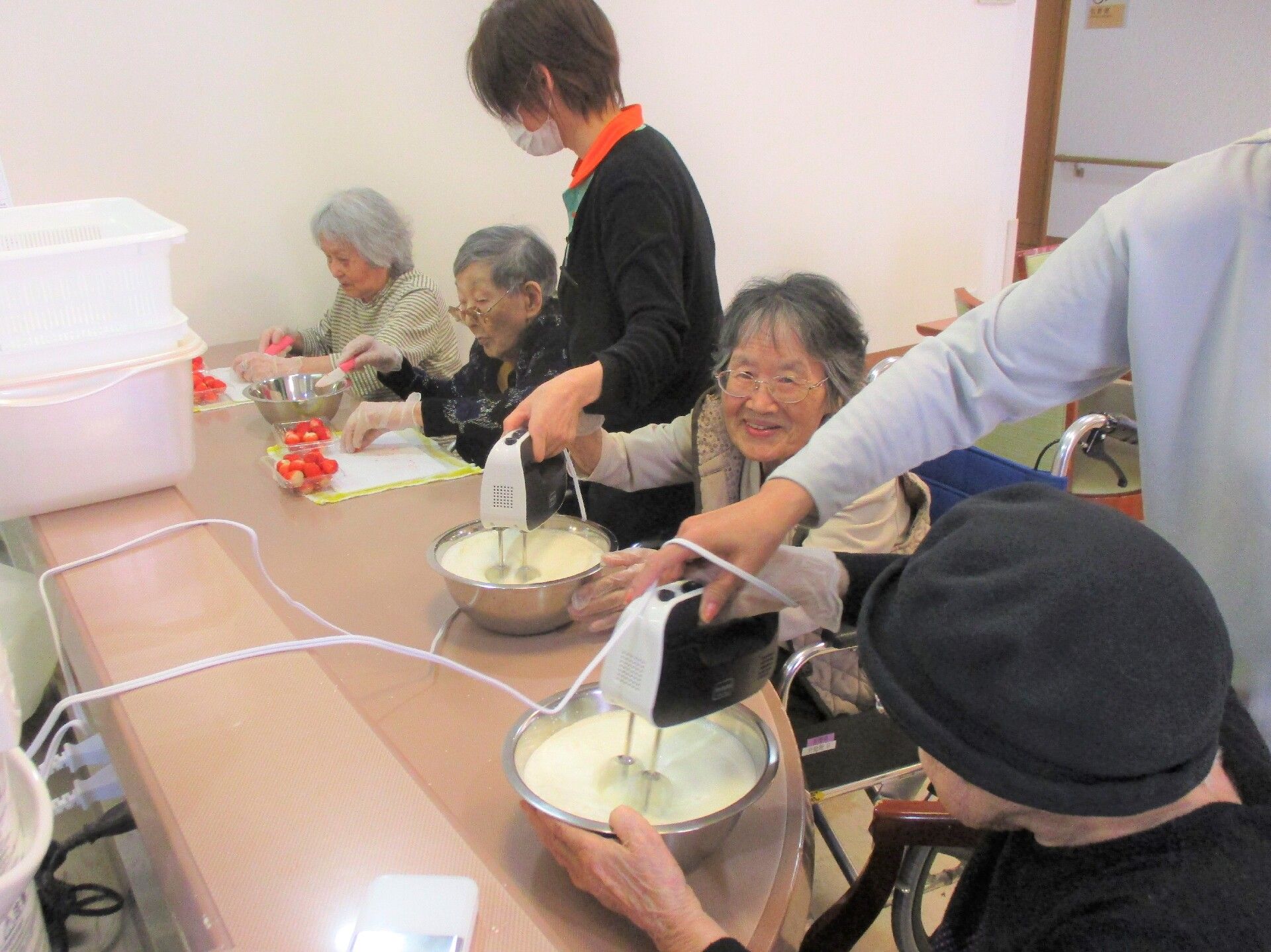 ボールとミキサーでおやつの材料をかき混ぜる高齢者と介助をするスタッフ