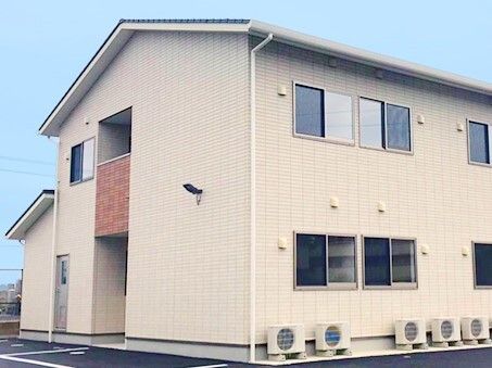 駐車場が付いた2階建ての建物。ベージュで家庭的な雰囲気が漂い、建物周辺には住宅もある。