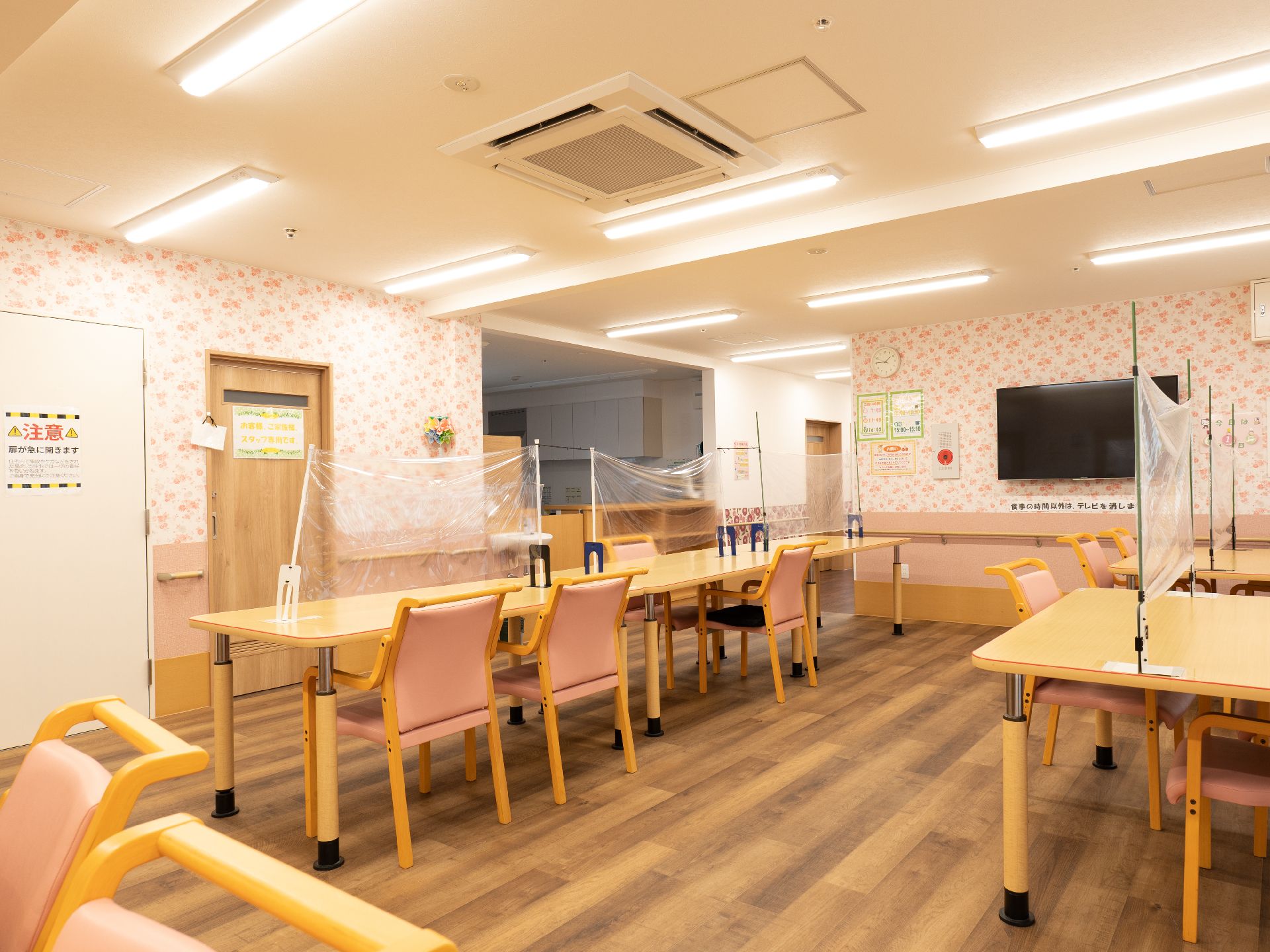 透明な仕切りが付いた木製の机が並べられたピンクの壁の広間