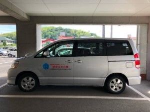 ポロシャツを着た男性スタッフが車いすを押し、高齢女性が誰かに向かって手を振っている。