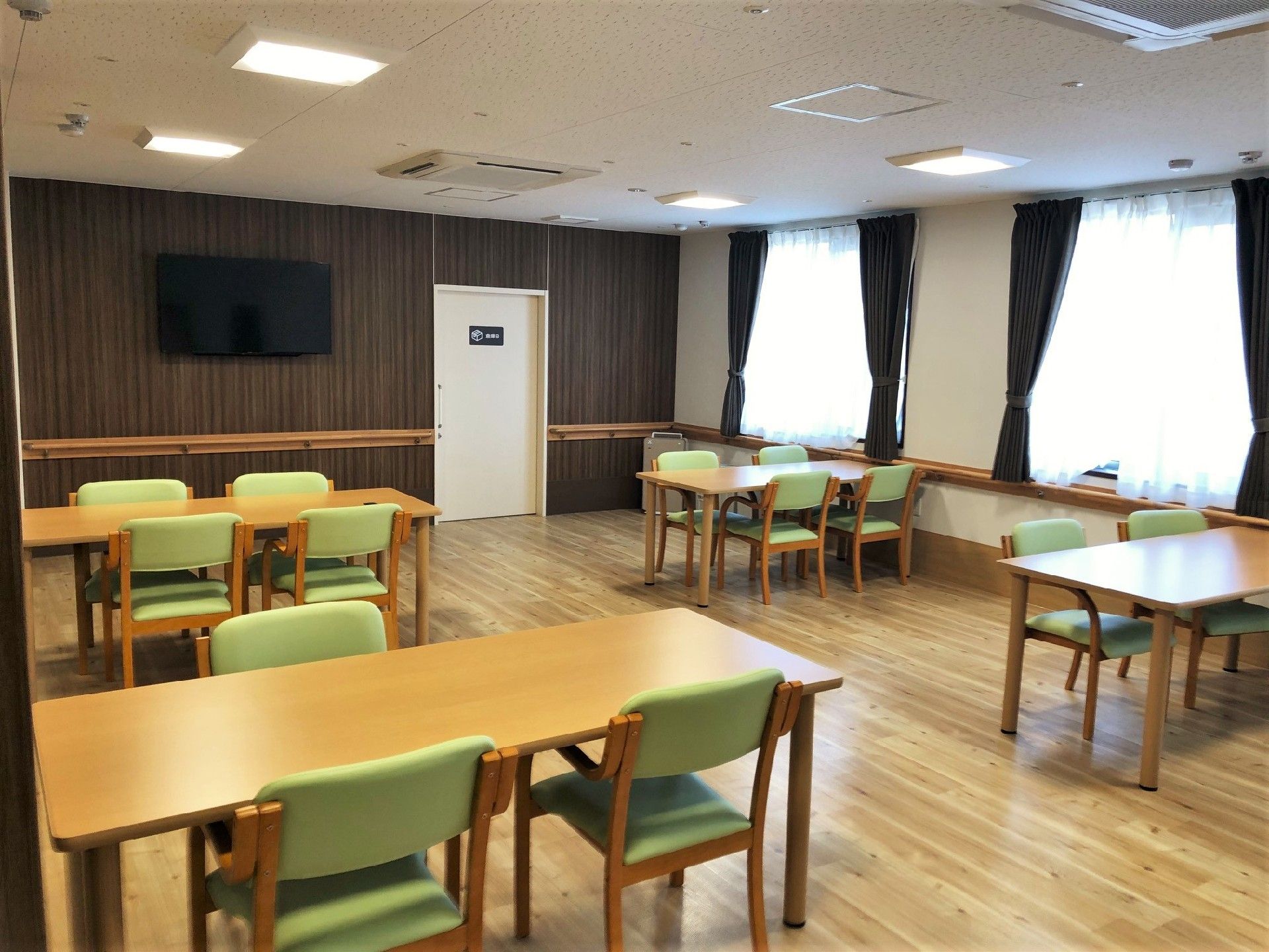 大型の壁掛けテレビが設置された食堂。４人掛けのダイニングテーブルが複数置かれ、大きな窓もたくさんある。