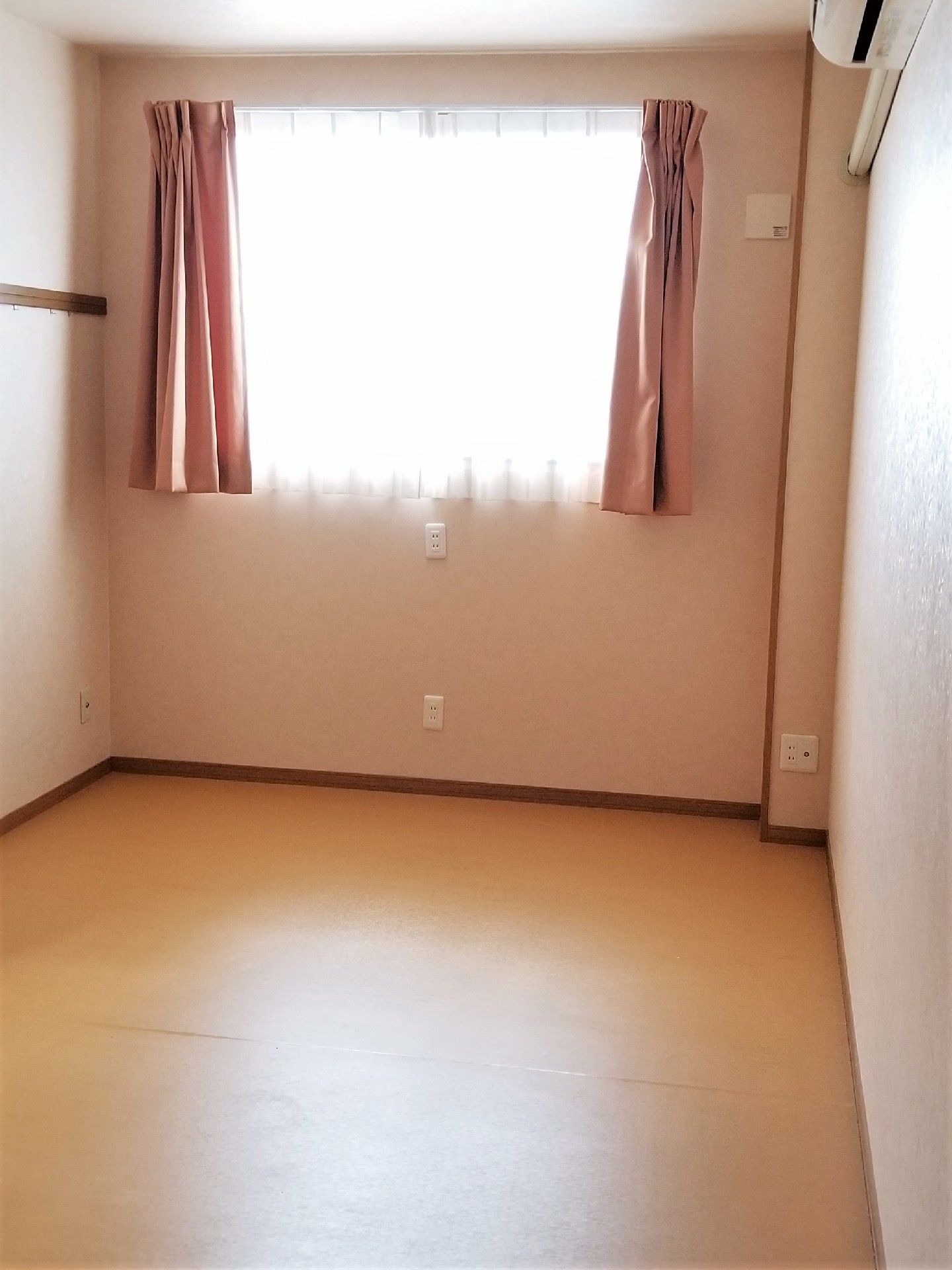 ピンクのカーテンが付いた。大き目の窓やエアコンが設置された、シンプルなフローリングの居室。