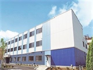 白をメインとした３階建ての建物。玄関には屋根が付き、周辺には緑もある。
