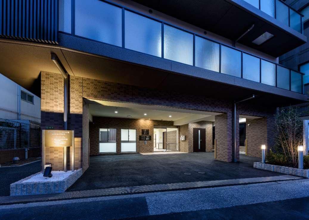 各フロアにベランダが付いた建物のエントランス部分。辺りが薄暗くなっていて、建物の中には明かりが灯っている。